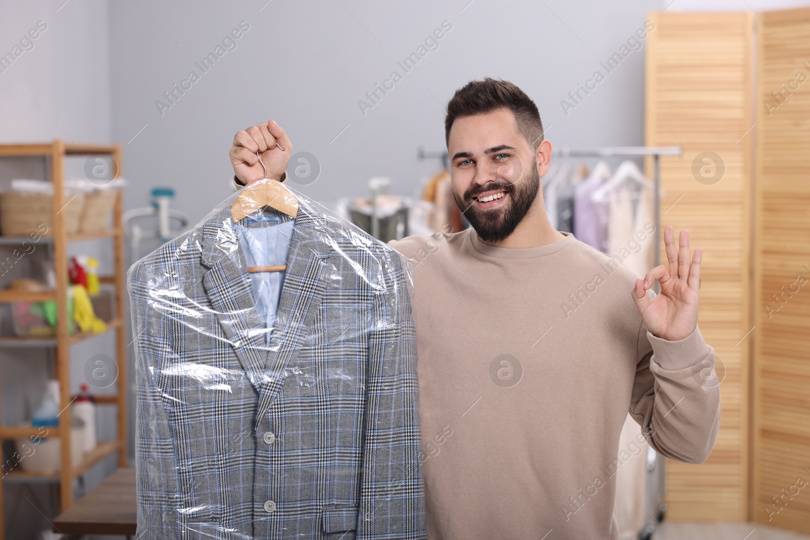 Photo of Dry-cleaning service. Happy man holding hanger with jacket in plastic bag and showing ok gesture indoors