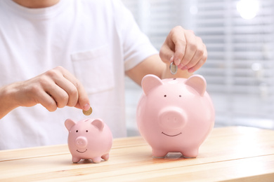 Photo of Man putting coins into piggy banks at wooden table, closeup