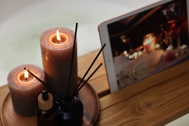 Photo of Wooden tray with tablet, burning candles and aroma products on bathtub, closeup