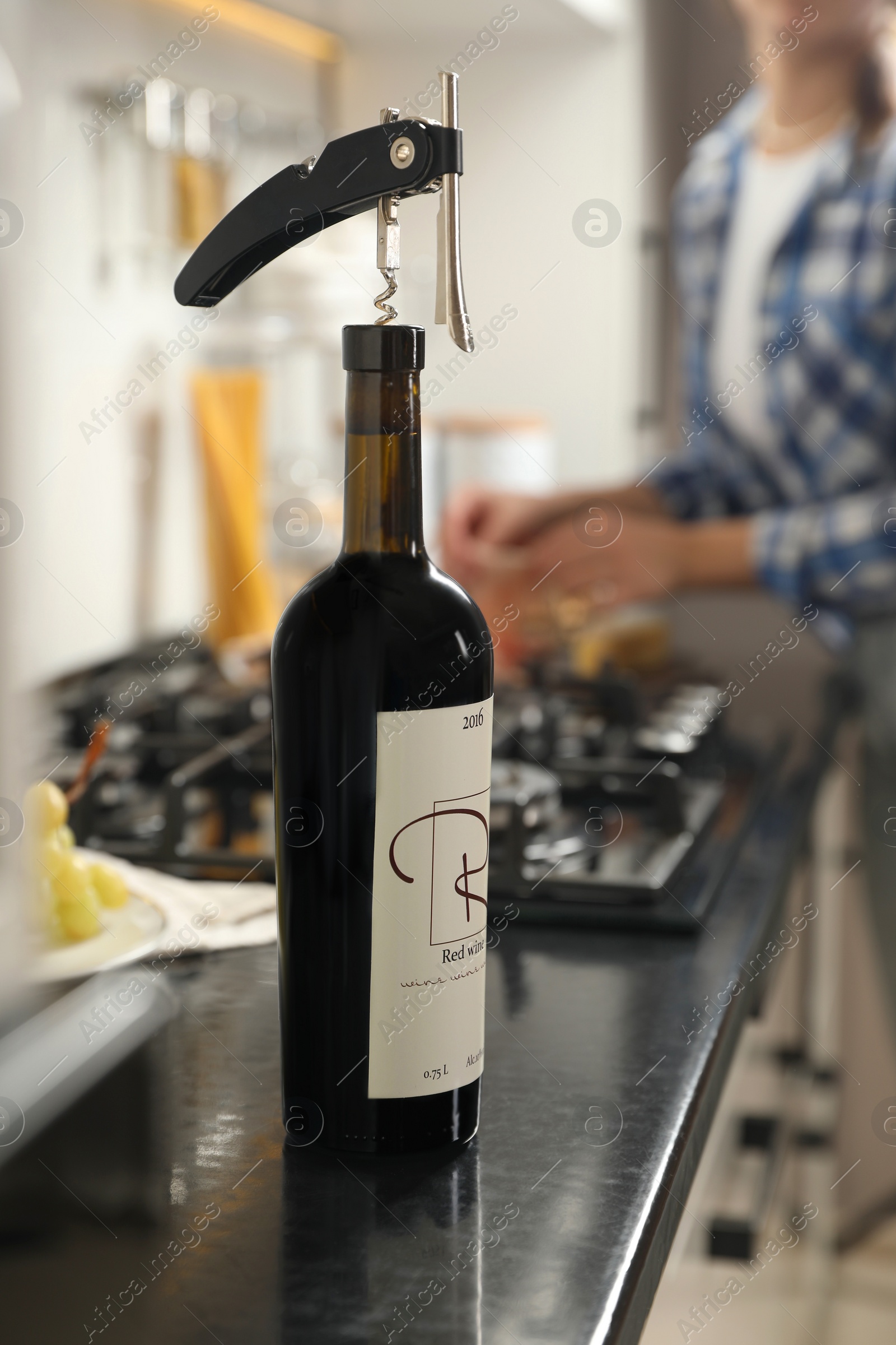 Photo of Wine bottle with corkscrew on black countertop, closeup. Woman in kitchen, selective focus