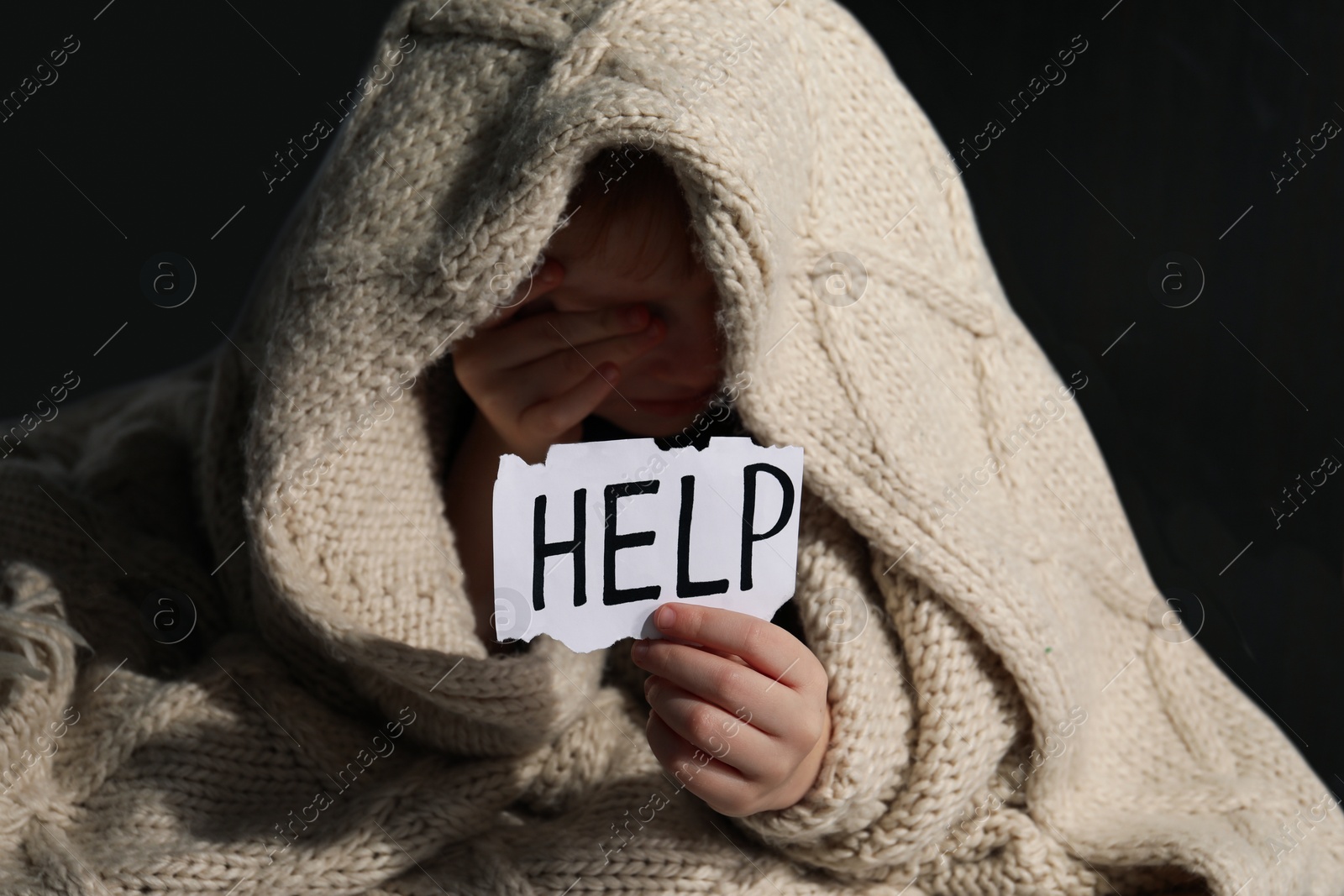 Photo of Scared little boy wrapped in blanket holding piece of paper with word Help indoors, focus on hand. Domestic violence concept