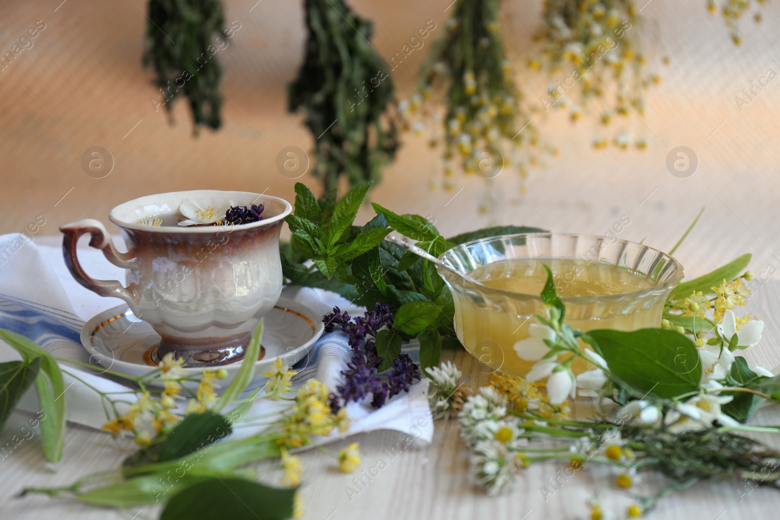 Photo of Cup of hot aromatic tea, honey and different fresh herbs on white wooden table