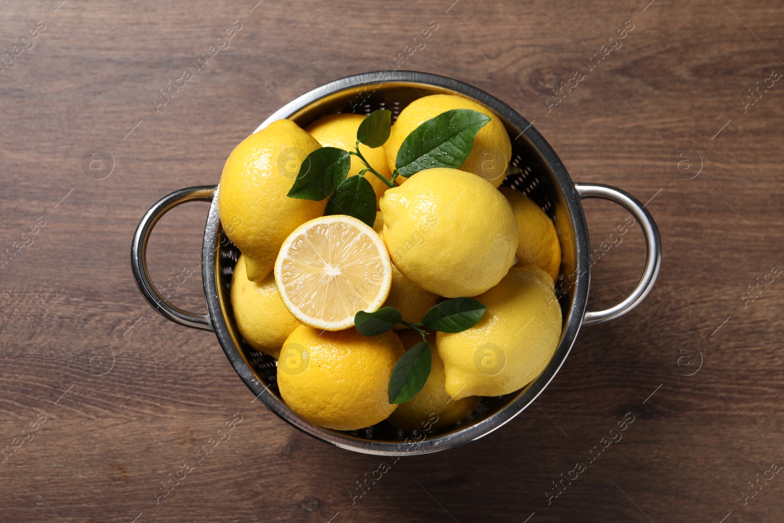 Photo of Fresh lemons and green leaves in colander on wooden table, top view