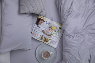 Photo of Cup of coffee and magazine on bed with soft silky bedclothes, top view