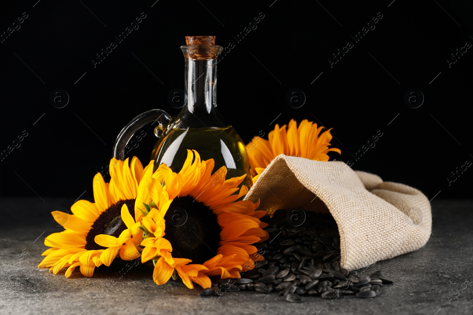 Photo of Sunflower cooking oil, seeds and beautiful flowers on grey table against black background