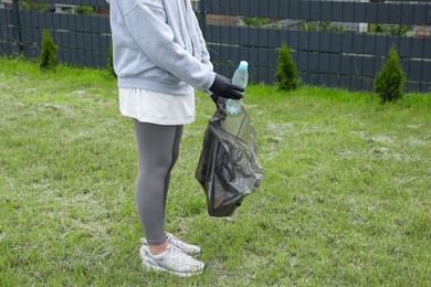 Woman with trash bag picking up plastic bottle outdoors, closeup. Recycling concept