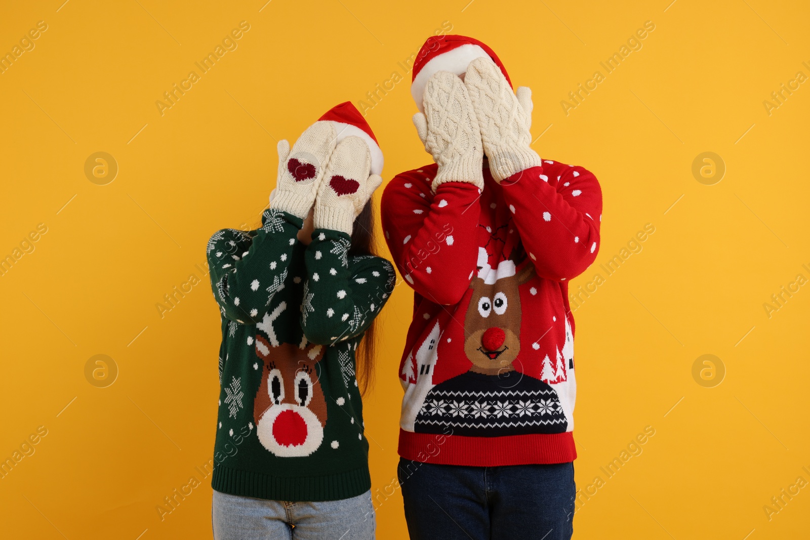 Photo of Couple in Christmas sweaters and Santa hats covering faces with hands in knitted mittens on orange background