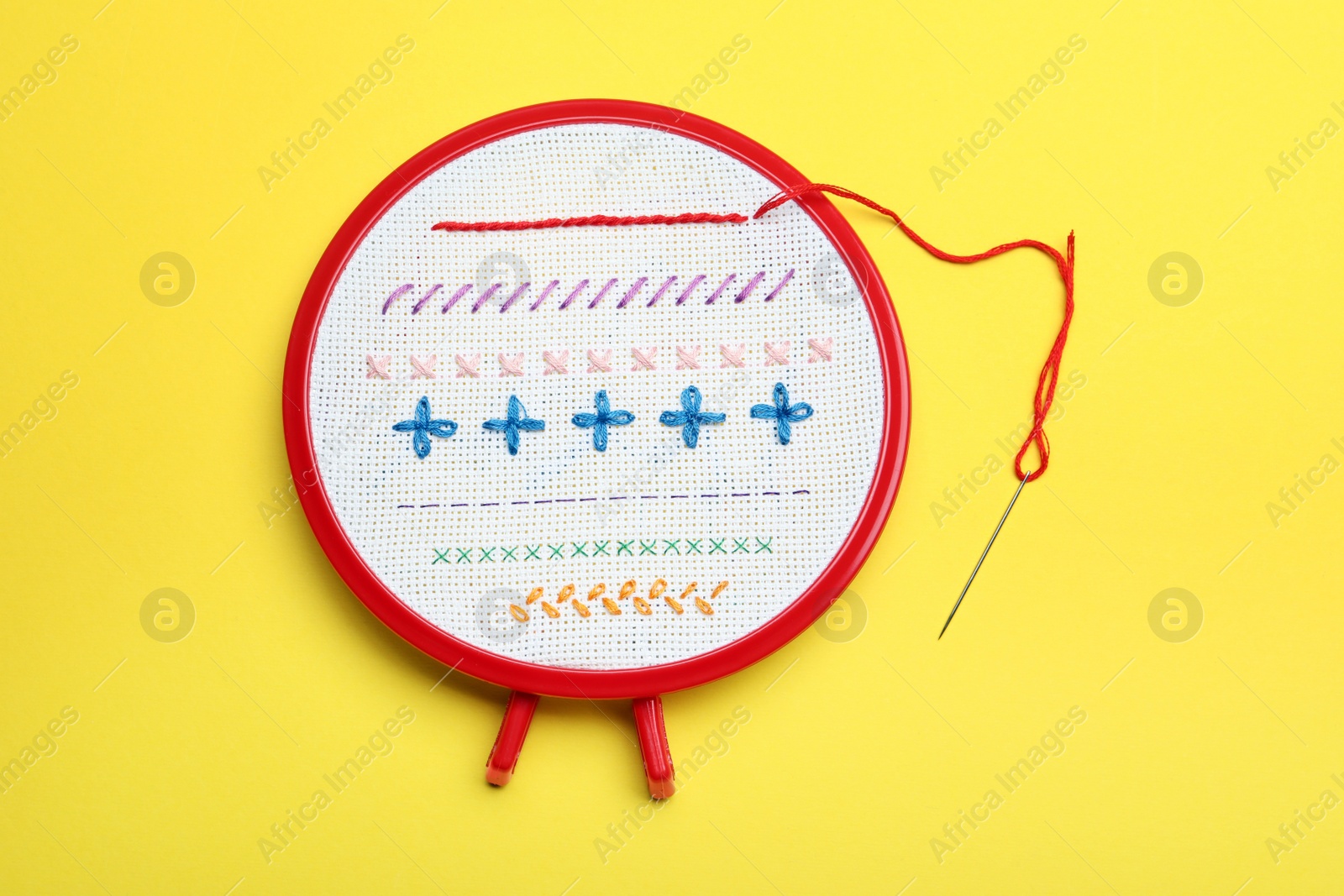 Photo of Fabric with colorful stitches in embroidery hoop and needle on yellow background, top view