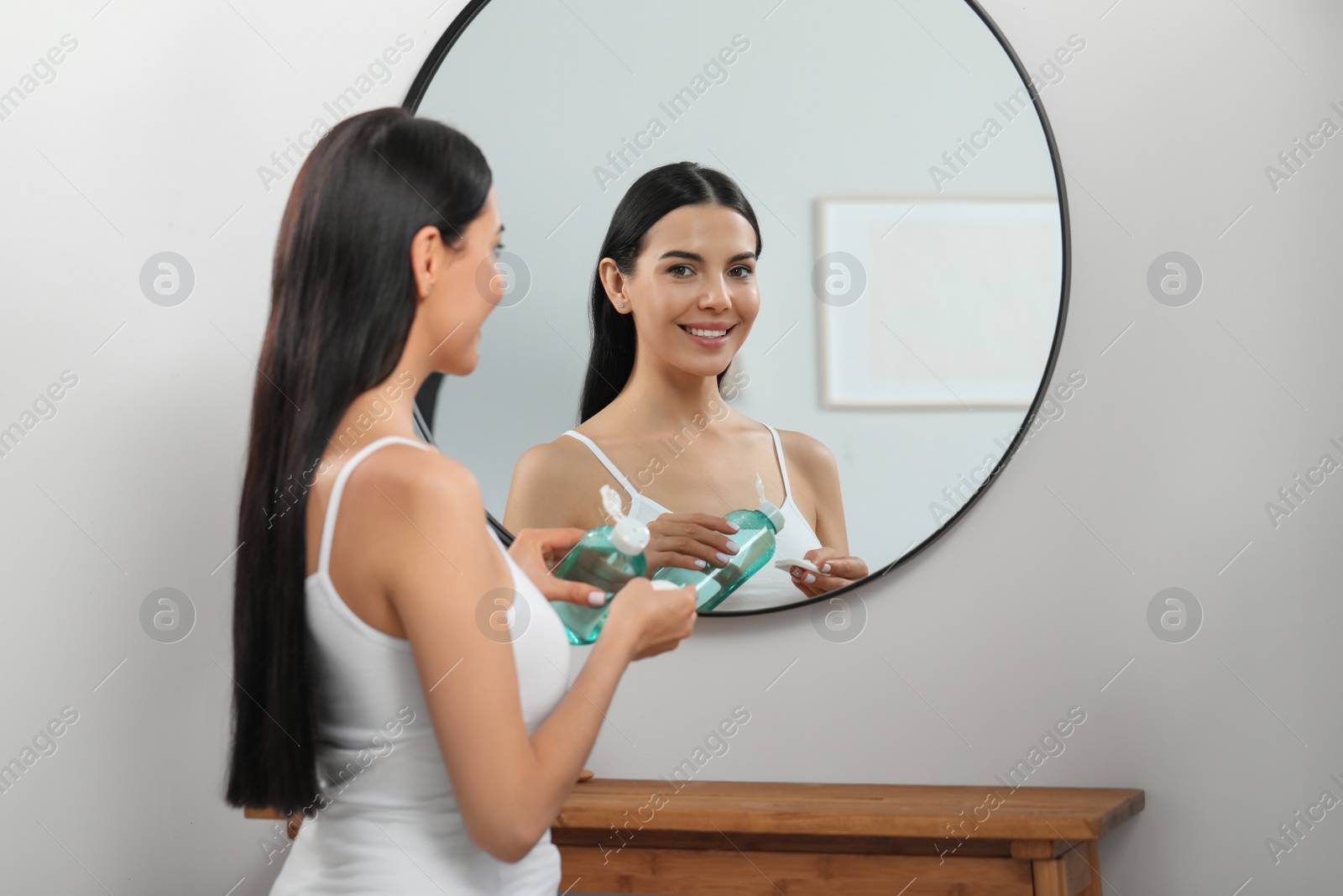 Photo of Young woman pouring micellar water onto cotton pad near mirror indoors