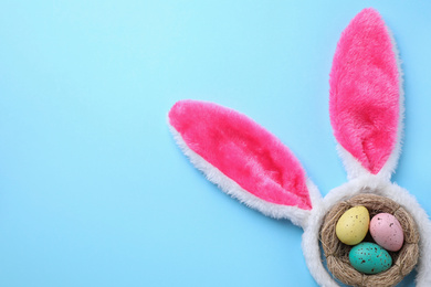 Headband with Easter bunny ears and dyed eggs in nest on light blue background, flat lay. Space for text