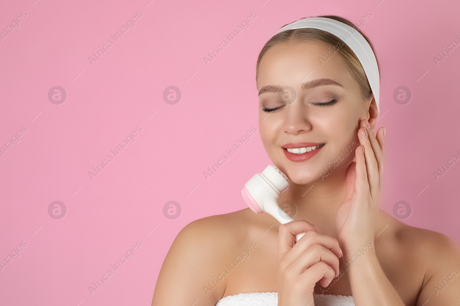 Photo of Young woman washing face with cleansing brush on pink background, space for text. Cosmetic product