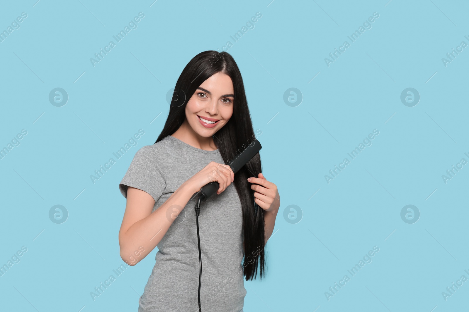 Photo of Beautiful happy woman using hair iron on light blue background. Space for text