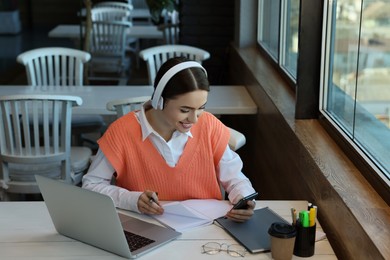 Young female student with laptop using smartphone while studying at table in cafe