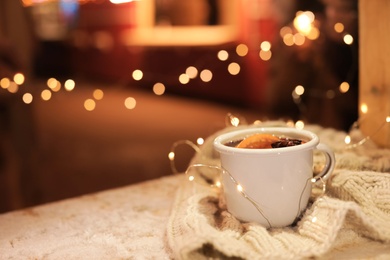 Cup of hot mulled wine and garland on table against blurred background. Space for text