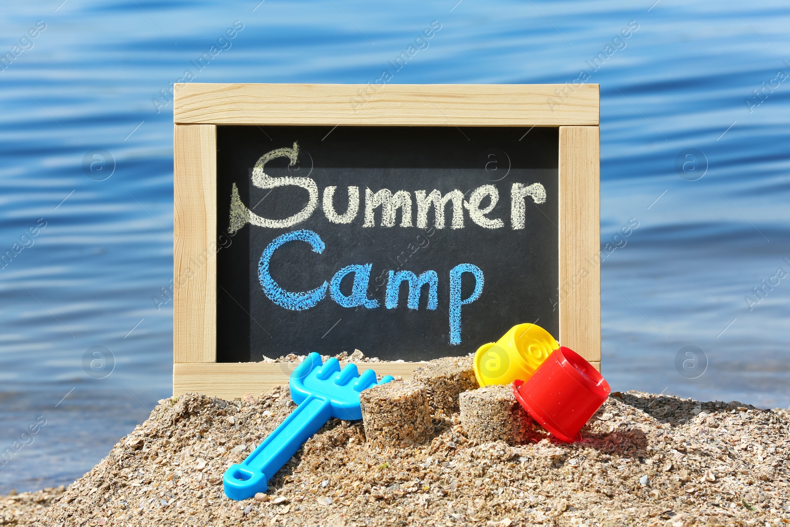 Photo of Little blackboard with text SUMMER CAMP and toys on sand near sea