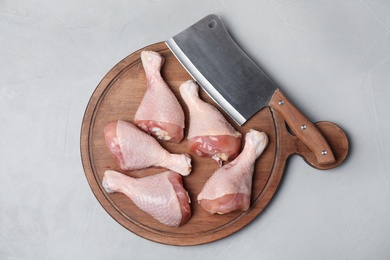 Photo of Wooden board with raw chicken drumsticks and butcher knife on gray background, top view. Fresh meat