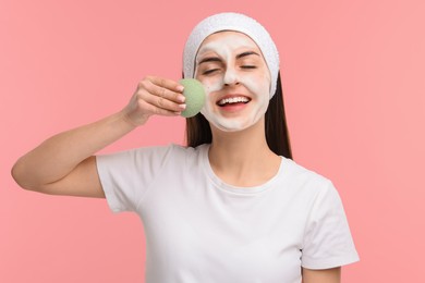 Photo of Young woman with headband washing her face using sponge on pink background