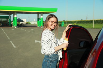 Photo of Beautiful young woman with hot dog opening car door at gas station. Space for text