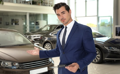 Photo of Young salesman with clipboard near car in modern dealership