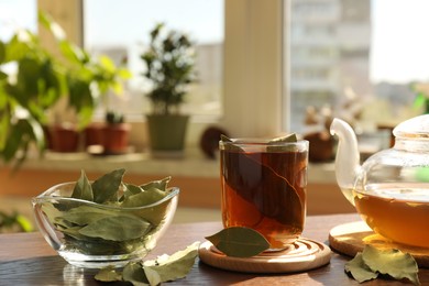 Photo of Cup of freshly brewed tea, bay leaves and teapot on wooden table indoors
