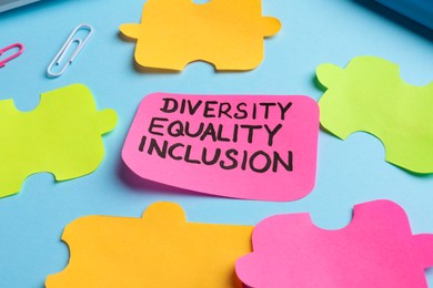 Photo of Sticky note with words Diversity, Equality, Inclusion and paper puzzle pieces on light blue background, closeup