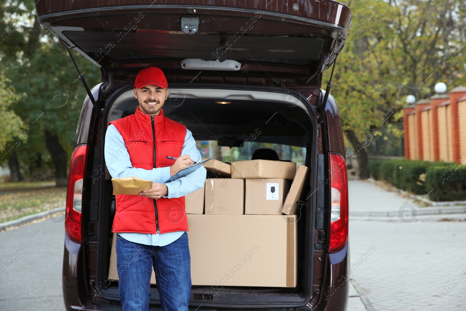 Photo of Young courier with parcel and clipboard near delivery van outdoors