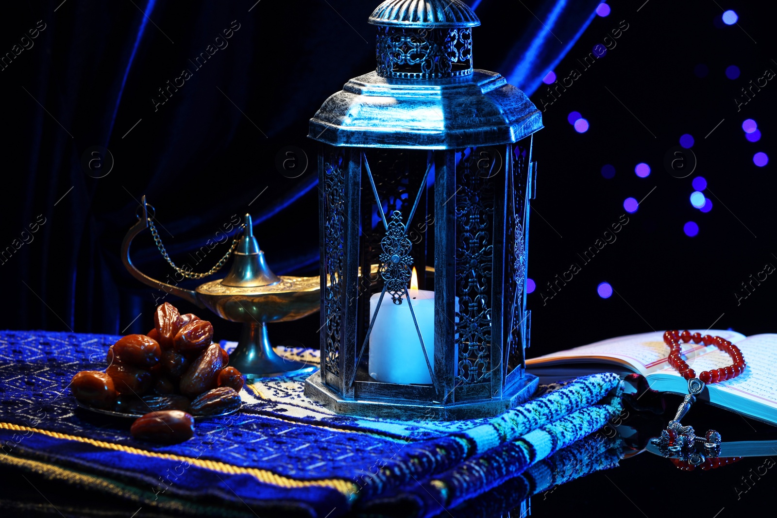 Photo of Arabic lantern, Quran, misbaha, Aladdin magic lamp, dates and folded prayer mat on mirror surface against blurred lights at night