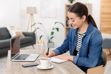 Photo of Young woman writing in notebook at wooden table indoors