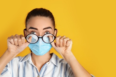 Woman wiping foggy glasses caused by wearing medical mask on yellow background. Space for text