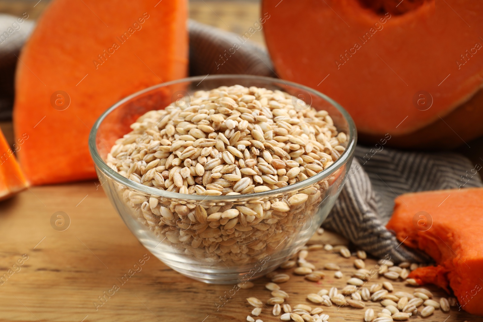 Photo of Dry pearl barley in bowl and pieces of pumpkin on wooden table, closeup