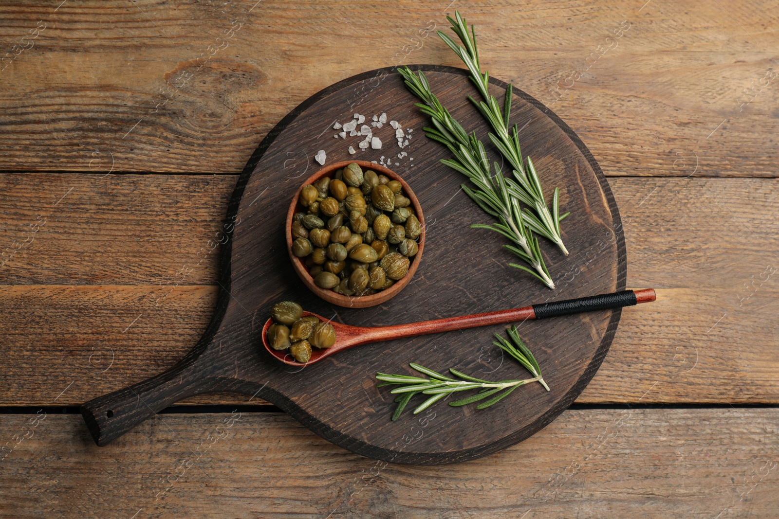 Photo of Capers, rosemary, salt and spoon on wooden table, top view