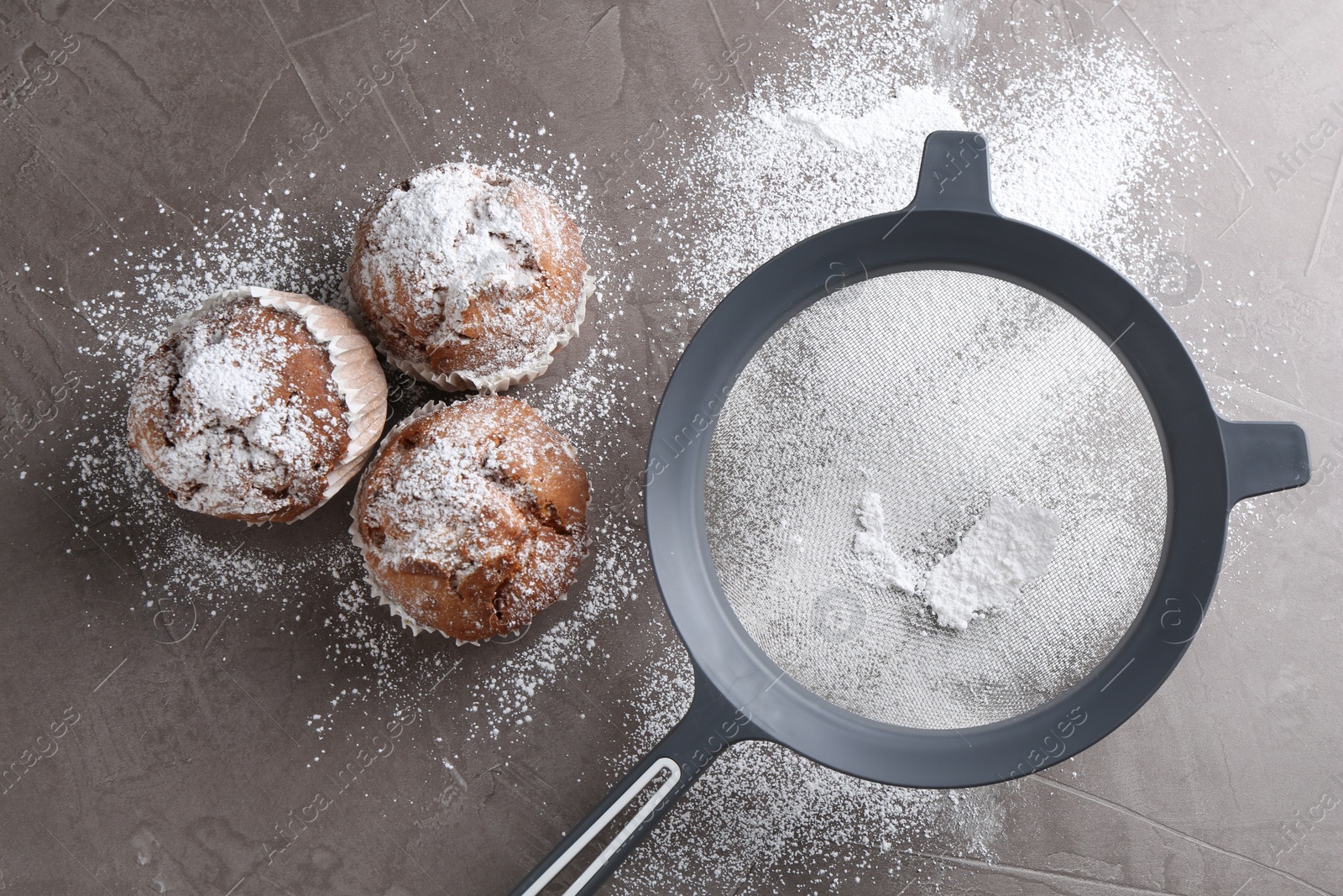 Photo of Sieve with sugar powder and muffins on grey textured table, flat lay