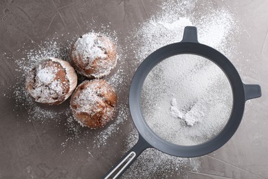 Sieve with sugar powder and muffins on grey textured table, flat lay