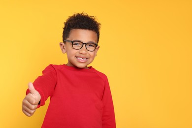 Photo of African-American boy with glasses showing thumb up on yellow background. Space for text