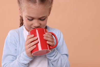 Photo of Cute girl drinking beverage from red ceramic mug on beige background, space for text