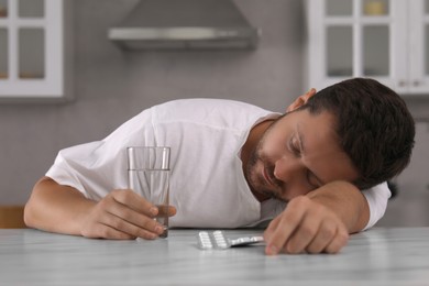 Man with glass of water and antidepressant pills sleeping at table in kitchen