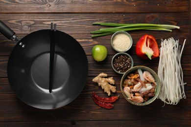 Black wok, chopsticks and products on wooden table, flat lay