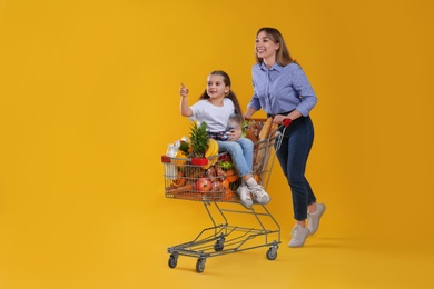 Mother and daughter with shopping cart full of groceries on yellow background