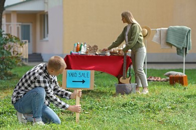 Man with sign Yard sale and woman near table of different items outdoors