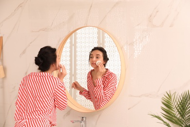 Photo of Young woman cleaning face with cotton pad near mirror in bathroom