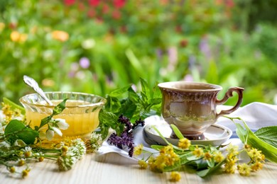 Photo of Cup of hot aromatic tea, honey and different fresh herbs on white wooden table outdoors