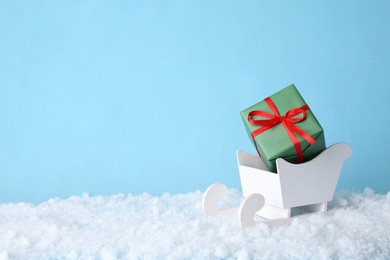 Photo of White wooden sleigh with Christmas gift box and artificial snow on light blue background, space for text