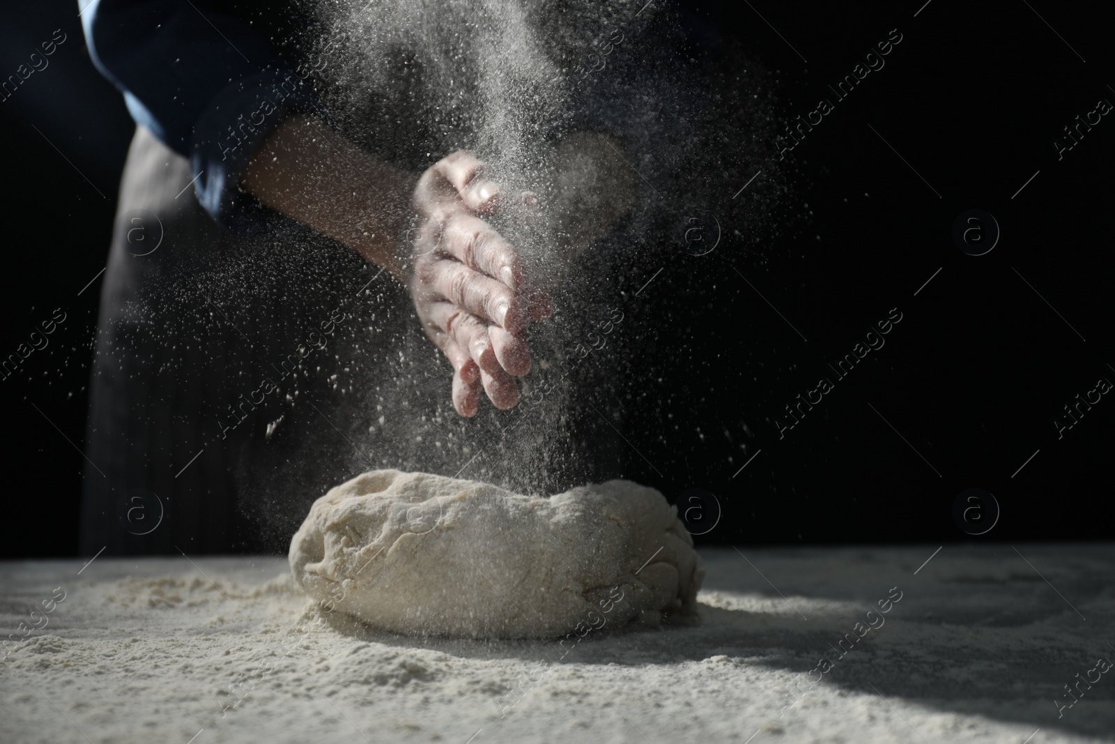 Photo of Making bread. Woman sprinkling flour over dough at table on dark background, closeup. Space for text