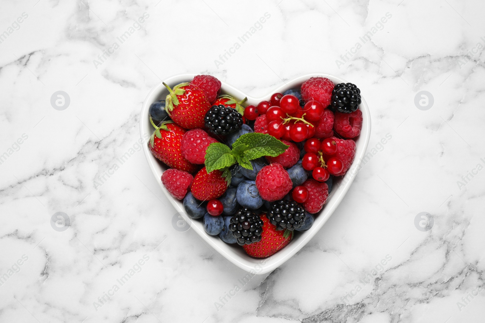 Photo of Mix of ripe berries in heart shaped bowl on white marble table, top view