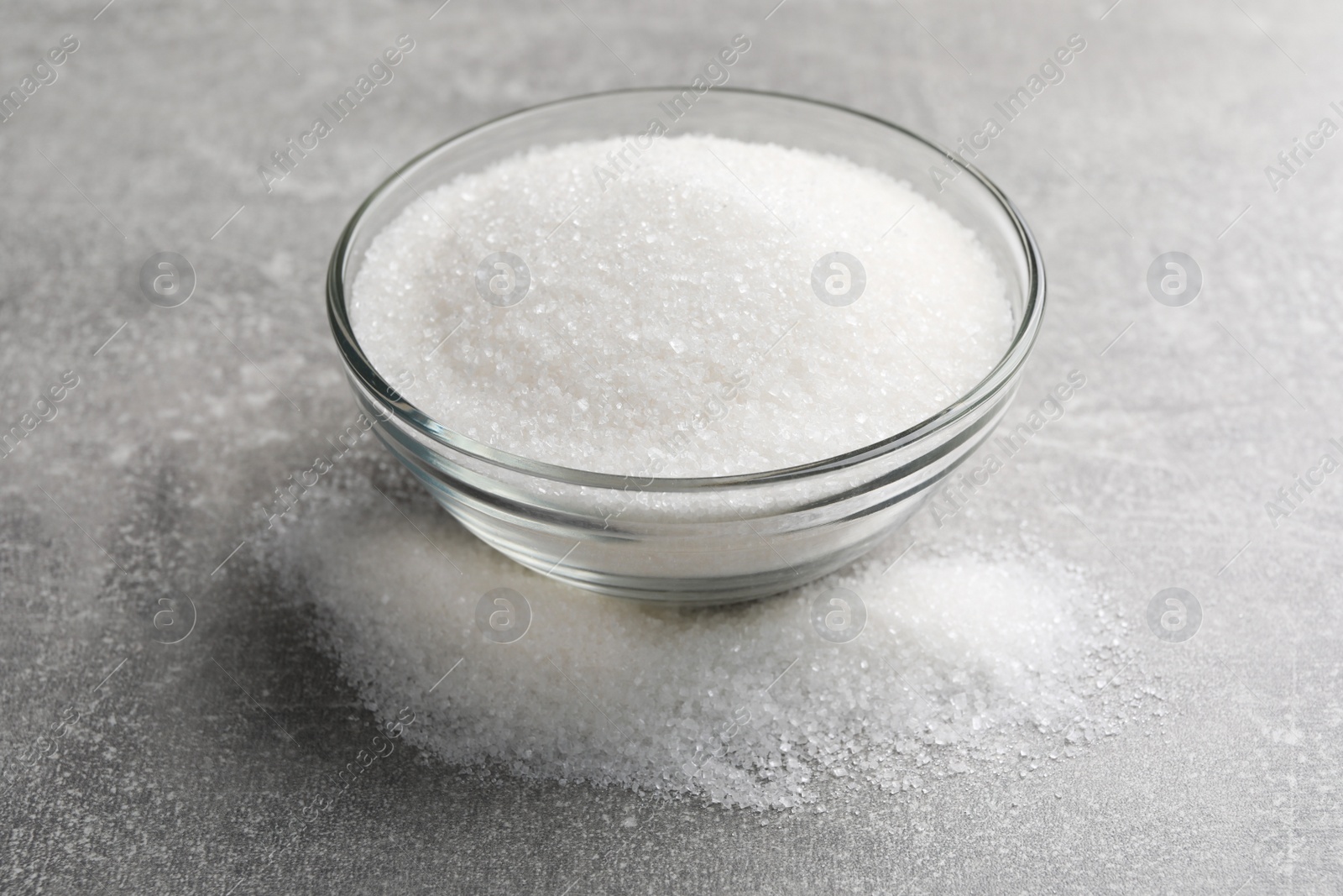 Photo of Granulated sugar and glass bowl on light grey table, closeup