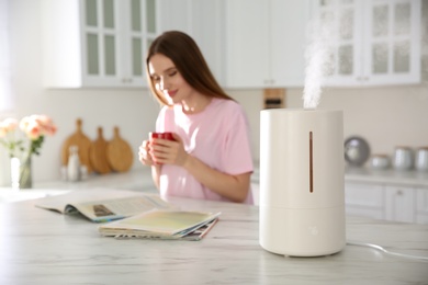 Photo of Modern air humidifier and blurred woman drinking coffee in kitchen