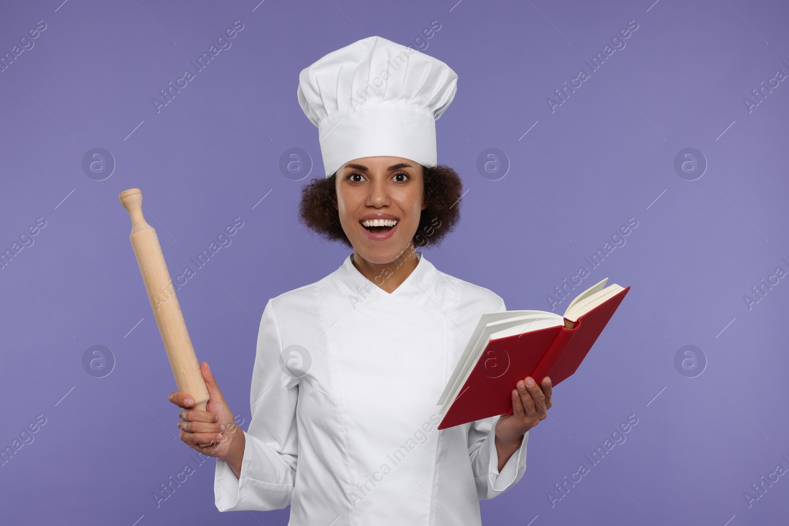 Photo of Emotional female chef in uniform holding rolling pin and recipe book on purple background