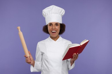 Photo of Emotional female chef in uniform holding rolling pin and recipe book on purple background