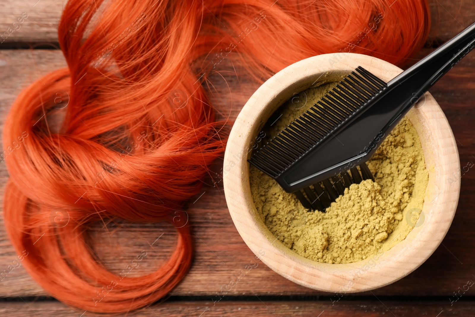 Photo of Bowl of henna powder, brush and red strand on wooden table, flat lay. Natural hair coloring