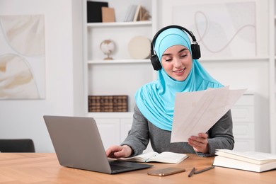 Muslim woman in hijab using laptop at wooden table in room